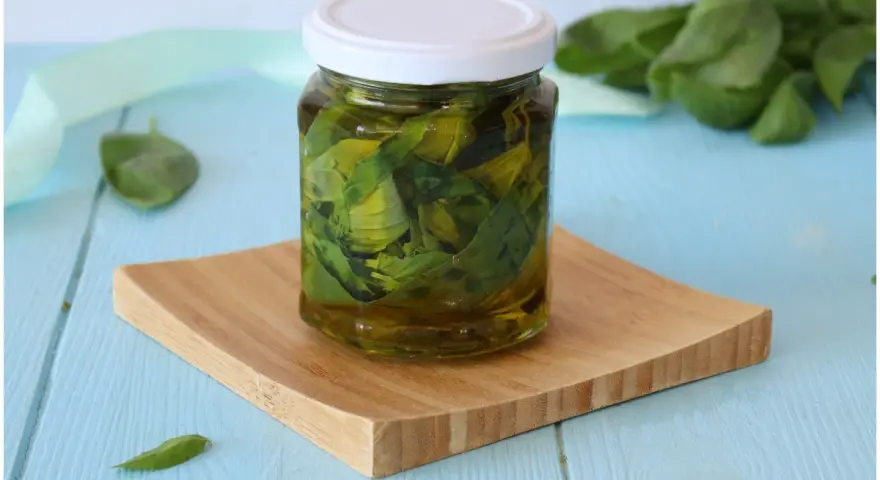 Un barattolo di olio al basilico fatto in casa, con foglie fresche immerse nell'olio dorato, su un tagliere di legno.