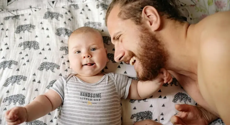Ein lächelnder Vater und sein Baby liegen auf einer Decke mit Bärenmuster. Die Frage, ob Autismus vererbbar ist, beschäftigt viele, und die Bindung zwischen Vater und Kind ist ein wichtiger Aspekt.