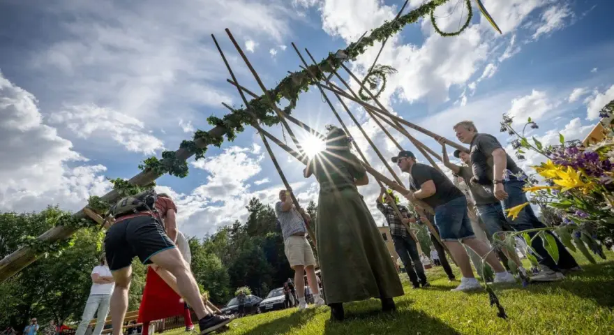 Midsommarstången reses av en grupp människor, en traditionell del av hur firar man midsommar. Solen skiner genom molnen.