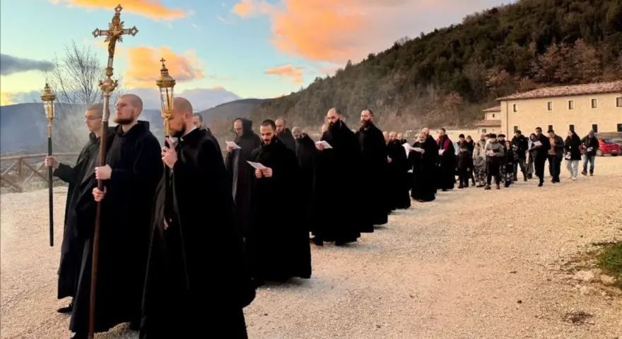 Monks in black robes process with a cross and lanterns, their solemn religious chants echoing as they walk towards a building at sunset.