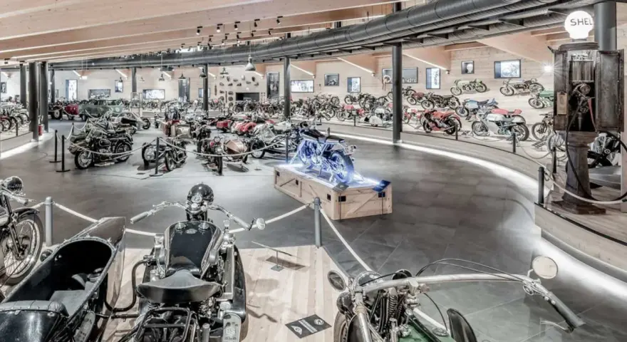 Wnętrze muzeum motocykli pełne zabytkowych maszyn, w tym motocykli z wózkami bocznymi i klasycznych samochodów.
