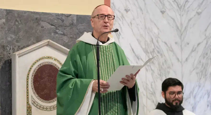 A man with a Benedictine title, wearing a green chasuble, speaks into a microphone while holding a paper.