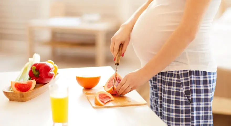 Schwanger, die Grapefruit für einen gesunden Snack schneidet. Nebenan liegen Paprika und ein Glas Saft.