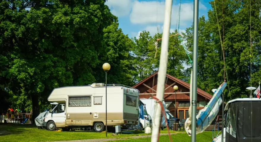 Słoneczny dzień na kempingu na Mazurach. Kamper stoi na trawie obok drewnianego budynku, w tle zielone drzewa i plac zabaw.