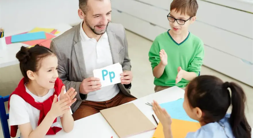 Nauczyciel pokazuje literę "Pp" podczas zebrania z rodzicami. Dzieci klaszczą, ucząc się alfabetu.