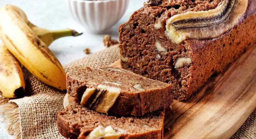 Saftiges, veganes Bananenbrot mit Walnüssen und Bananenscheiben, daneben frische Bananen und eine Schüssel Nüsse.