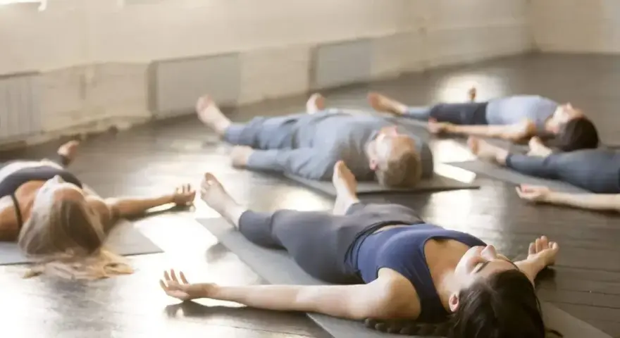 Grupa osób leży na matach w pozycji Savasana, wykonując ćwiczenia na uspokojenie.