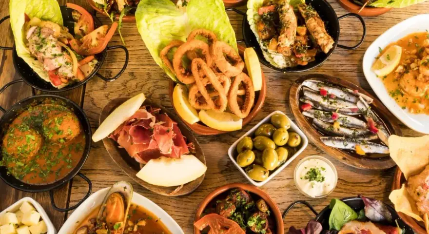 Tapas selber machen: Eine Auswahl an köstlichen spanischen Kleinigkeiten wie frittierte Tintenfischringe, Sardinen, Oliven und Schinken auf einem Holztisch.
