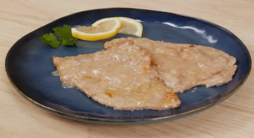 Due scaloppine al limone cremose, guarnite con prezzemolo e spicchi di limone, su un piatto blu.