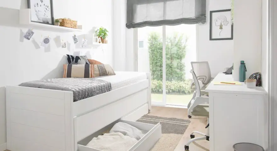 Habitación estrecha y alargada con cama blanca, escritorio y silla. Ventana grande con vistas al jardín.