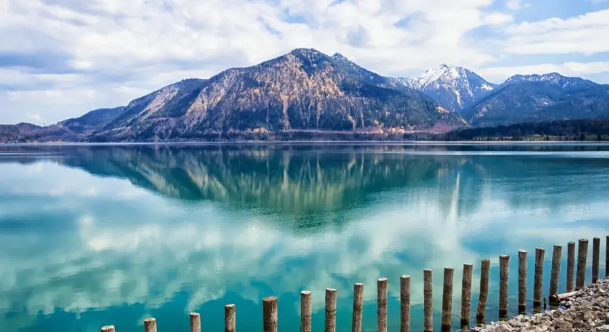Spiegelglatter See mit Bergen im Hintergrund, wie man ihn in Bayern sehen kann. Holzpfähle ragen aus dem türkisfarbenen Wasser.