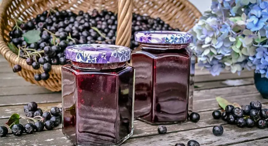 Zwei Gläser mit Aronia-Marmelade, bereit zum Verarbeiten. Ein Korb voller Aronia-Beeren steht im Hintergrund.