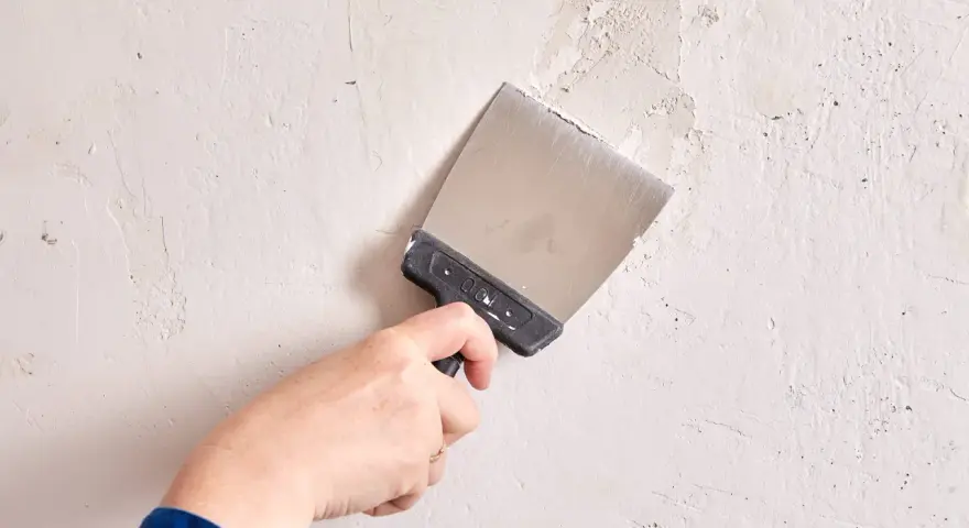 Mano con espátula metálica alisando una pared de yeso con textura irregular.