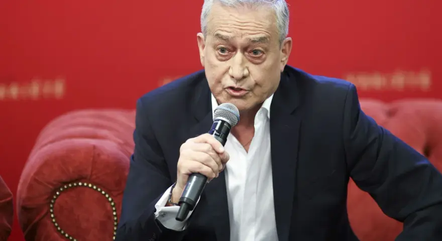 Janusz Szydłowski przemawia do mikrofonu, siedząc na czerwonej, pikowanej kanapie.