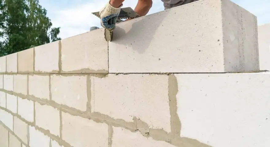Budowanie ściany z bloczków betonowych. Ręka w rękawicy nakłada zaprawę na kolejny element.