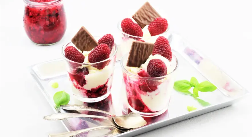 Drei Gläser mit cremigem Dessert, Himbeeren und Schokolade. Ein Glas mit Himbeermarmelade daneben. Schnelles Eis für den Sommer.