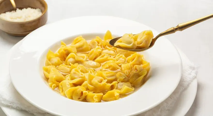 Un cucchiaio d'oro solleva un boccone di cappelletti in brodo fumante da un piatto bianco.