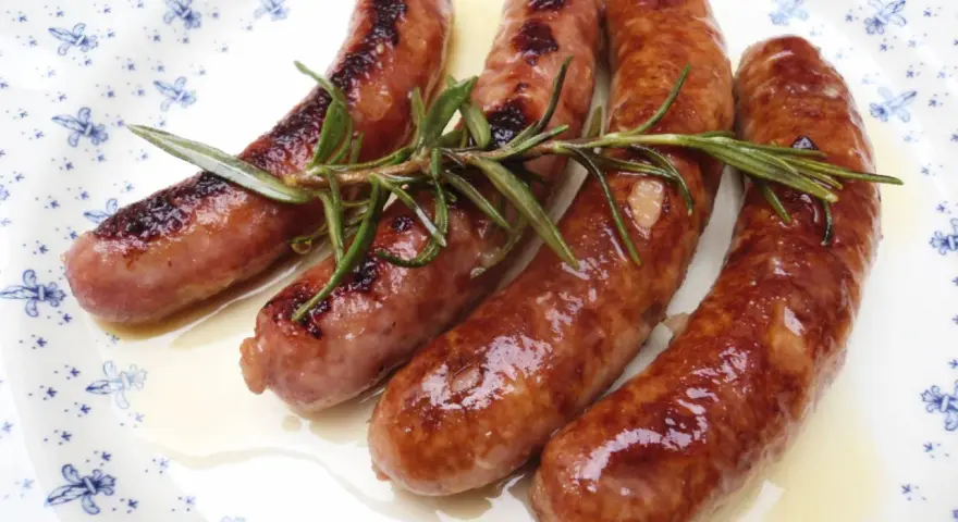 Salchichas al vino, doradas y jugosas, reposan sobre un plato blanco con delicados motivos azules, adornadas con ramitas de romero fresco.
