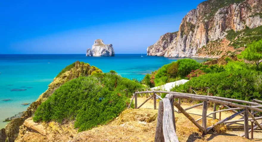 Sentiero con staccionata in legno conduce a una spiaggia con acqua cristallina e l'isolotto Pan di Zucchero in Sardegna.