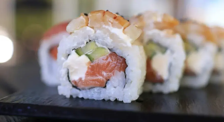 Uramaki co to? Rolka sushi z łososiem, awokado i krewetką, ryż na zewnątrz, na czarnej desce.