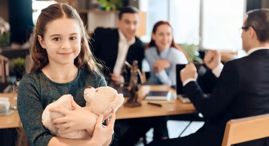 Uśmiechnięta dziewczynka z misiem w ramionach, podczas gdy rodzice i prawnik omawiają zmianę nazwiska dziecka.