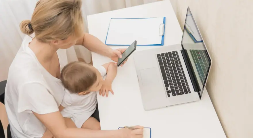 Kobieta z dzieckiem na kolanach sprawdza na telefonie informacje o zasiłku macierzyńskim 80 czy 100% i 60. Obok laptop i notatnik.