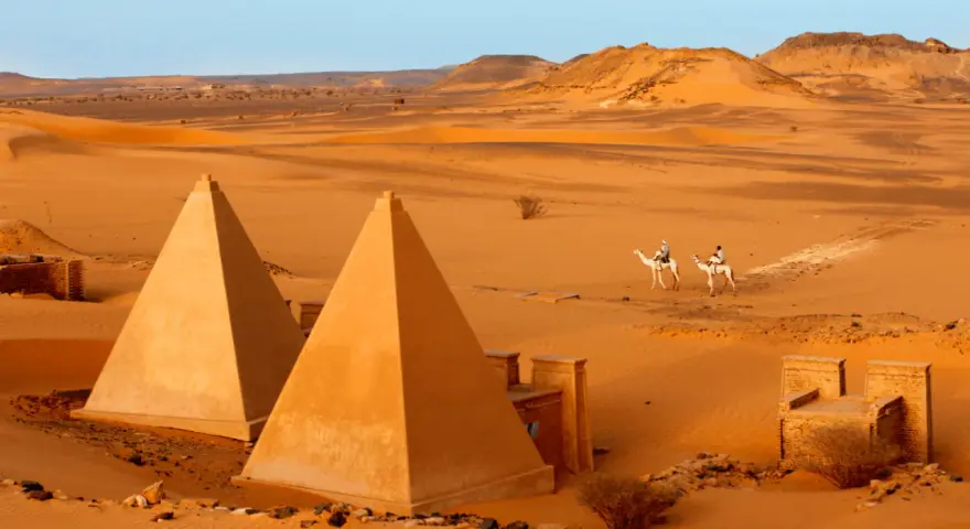 Pustynny krajobraz Sudanu z piramidami i dwoma podróżnikami na wielbłądach.