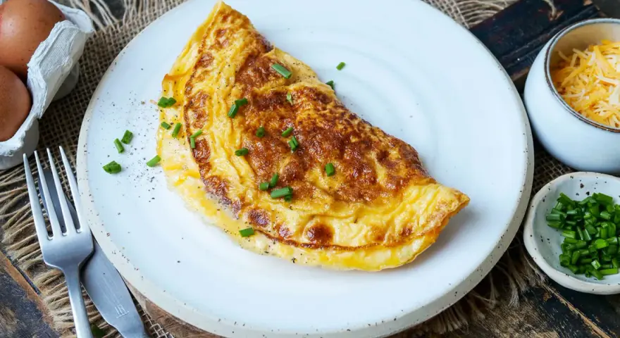 Goldbraunes Omelett mit Käse und Schnittlauch, perfekt für ein schnelles Omelette Rezept. Daneben Eier, Käse und Schnittlauch.