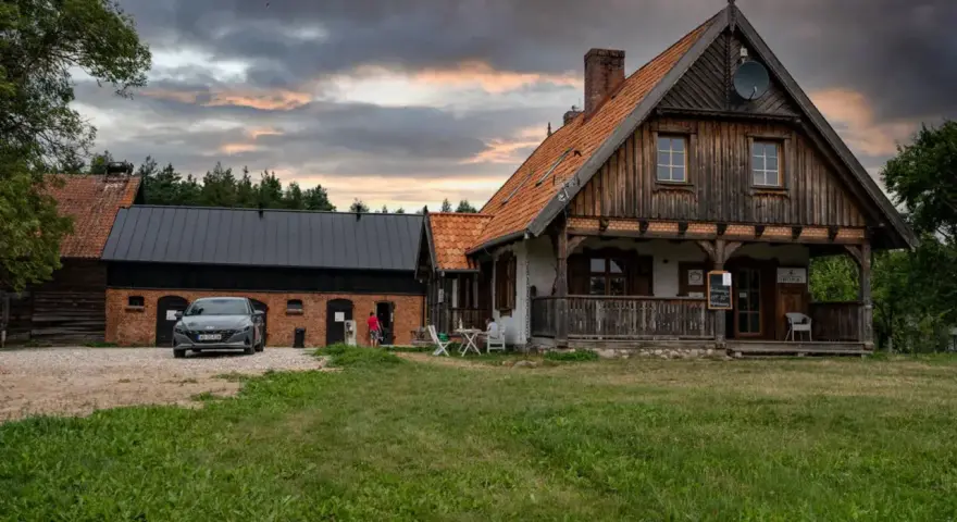 Szelągówka camping: drewniane domki i budynki gospodarcze, samochód na żwirowym podjeździe, zachmurzone niebo.