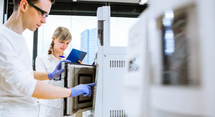 W laboratorium młoda para naukowców pracuje przy aparaturze. On w okularach, ona z notatkami.