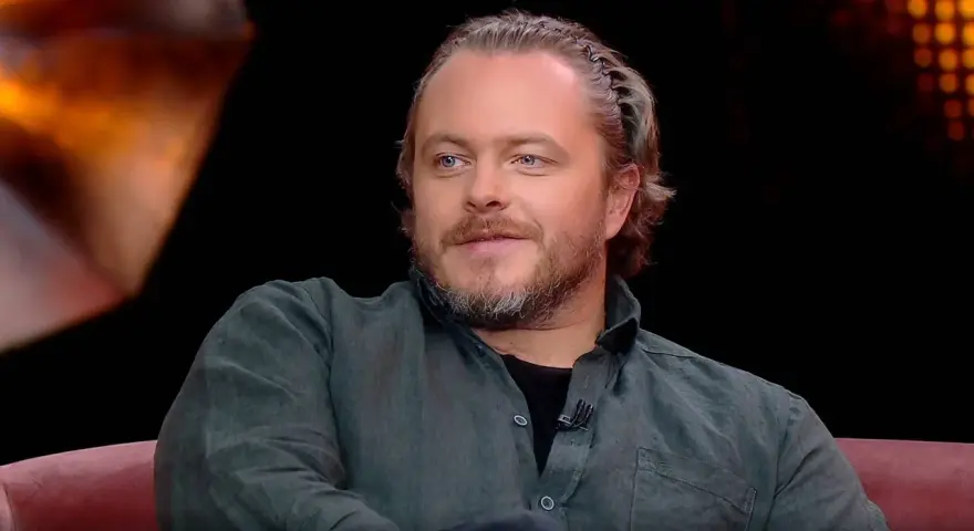 Tomasz Schuchardt, znany z wielu filmów, seriali i programów, z charakterystyczną fryzurą i niebieskimi oczami, siedzi na kanapie.