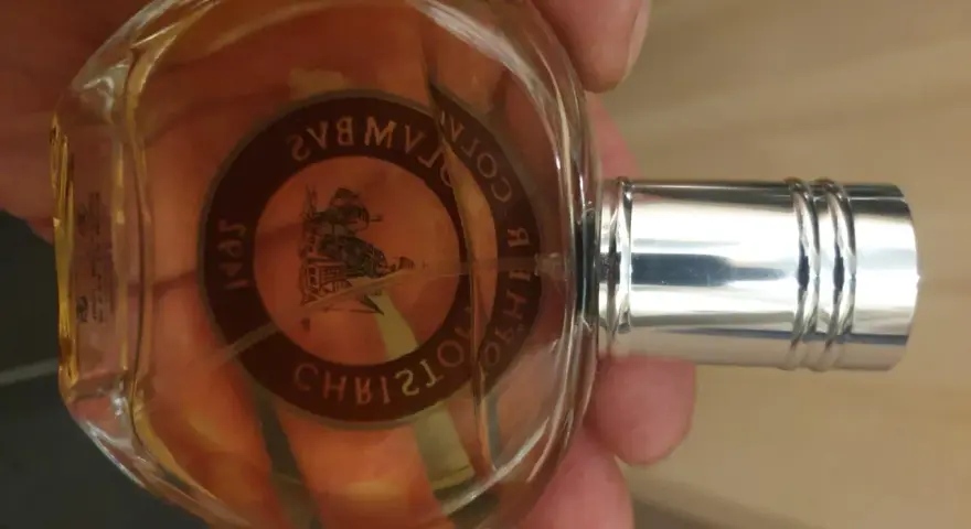 Szklana butelka z bursztynowym płynem, ozdobiona okrągłą etykietą z wizerunkiem statku i napisem "Christopher Columbus perfumy".