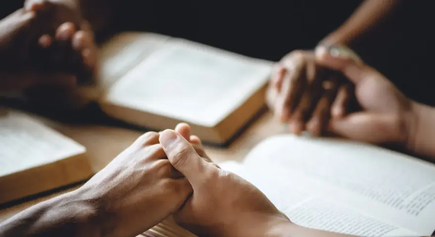 Hände halten sich über aufgeschlagenen Bibeln, ein Zeichen dafür, wie beten Christen: in Gemeinschaft, mit Glauben und Hoffnung.