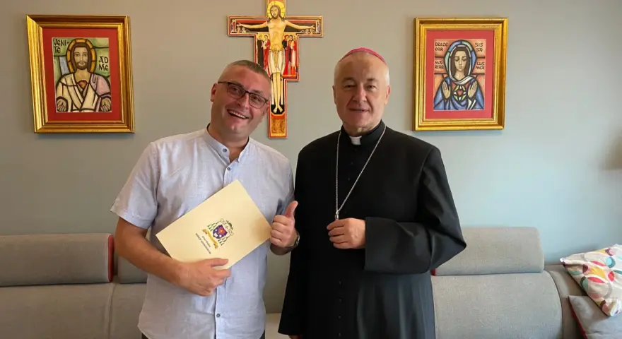 Uśmiechnięty mężczyzna z dokumentem i biskup pozują na tle krzyża i obrazów. Mężczyzna trzyma dokument z herbem, a biskup ma na sobie sutannę.