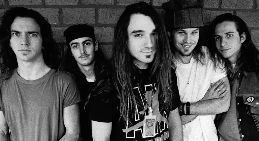 Cinq membres du groupe Pearl Jam posent devant un mur de briques. L'un d'eux porte un t-shirt avec le logo du groupe.