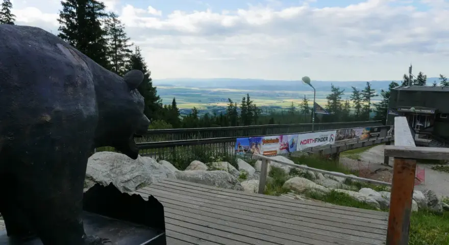 Pomnik niedźwiedzia na tle panoramy Tatr poza szlakiem. Drewniana kładka prowadzi do budynku z widokiem na dolinę.