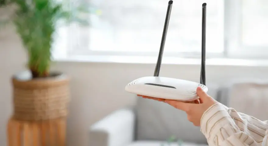 Dłoń trzyma biały router z dwiema antenkami. Dowiedz się, jak zresetować router, aby rozwiązać problemy z siecią.