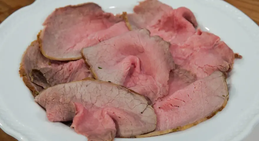 Fette sottili di arrosto di manzo rosa e succulento, servite su un piatto bianco.