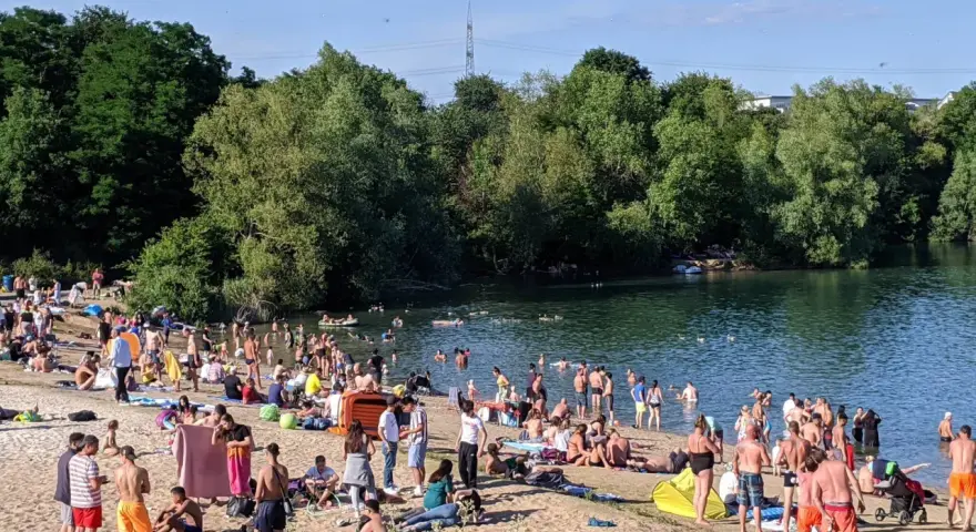 Ein sonniger Tag am **Badesee Bonn**. Viele Menschen genießen das Wasser und den Strand, umgeben von grünen Bäumen.