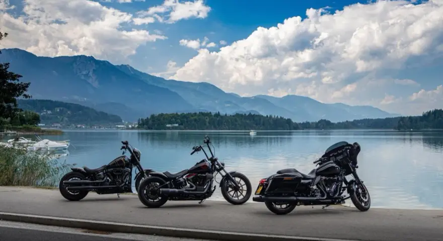 Tre moto parcheggiate sul lungolago, pronte per il faaker see motoraduno. Montagne sullo sfondo, cielo azzurro con nuvole bianche.