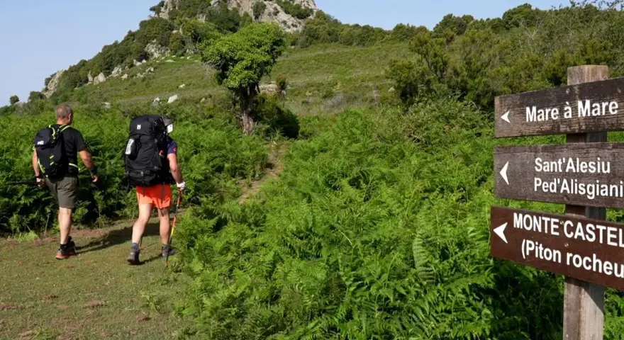 Deux randonneurs suivent un sentier bordé de fougères. Un panneau indique la direction de Mare a Mare Nord.