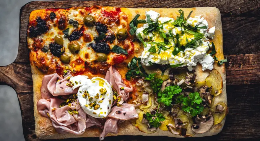 Vier verschiedene Beläge auf einer rechteckigen pizza al taglio: Oliven, Ricotta mit Kräutern, Mortadella mit Burrata und Kartoffeln mit Pilzen.