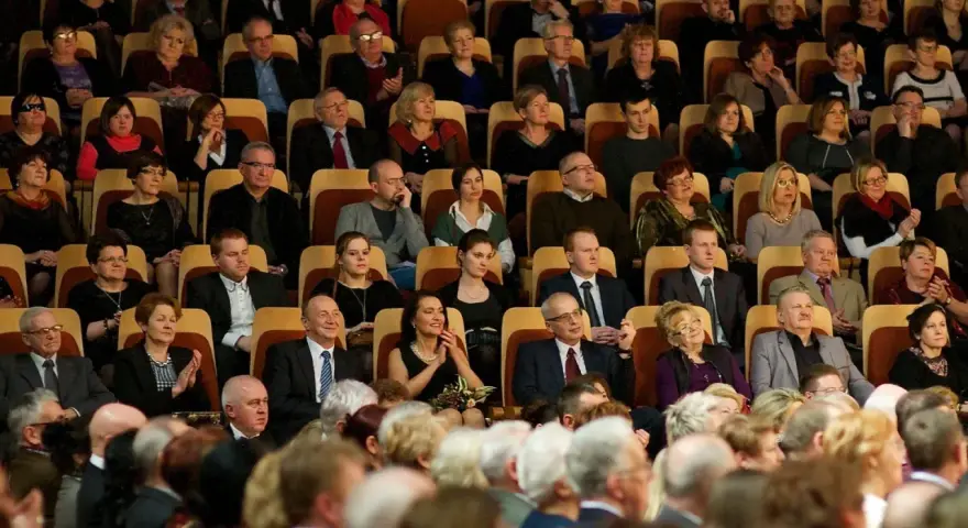 Elegancko ubrani ludzie siedzą w filharmonii. Panie w sukienkach, panowie w garniturach. To inspiracja, jak się ubrać do filharmonii.