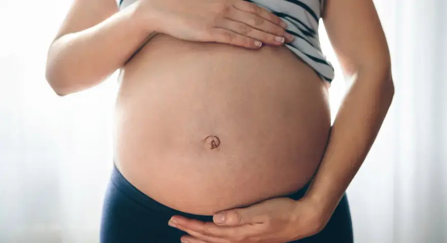 Brzuszek w ciąży z widocznym pępkiem, otoczony dłońmi przyszłej mamy.