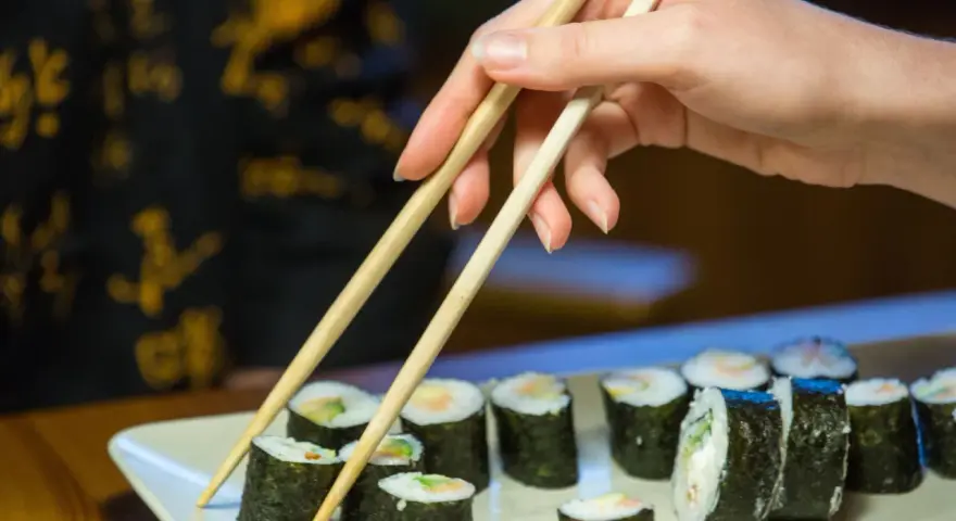 Dłoń z pałeczkami sięga po rolkę sushi. Na talerzu ułożone są kawałki sushi, każdy z nich to mała porcja smaku i kalorii.