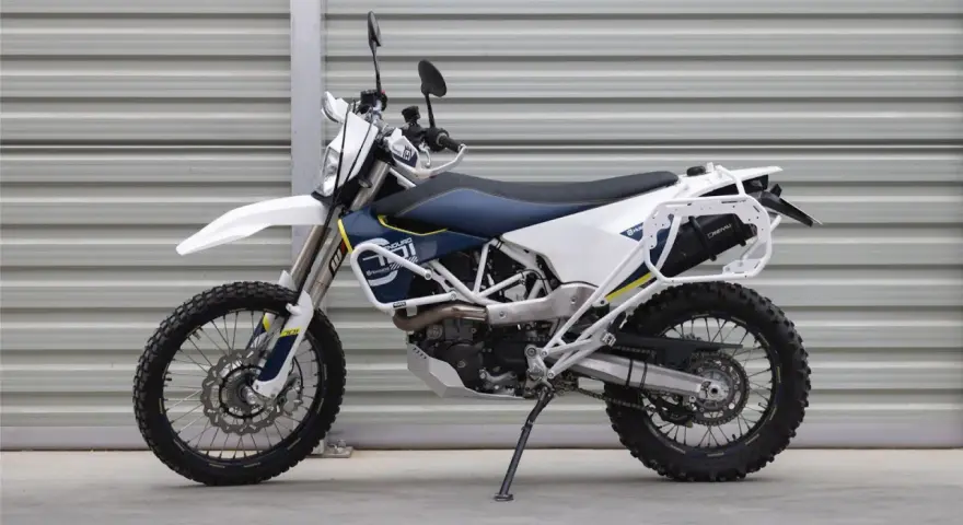 Biało-niebieski motocykl Husqvarna z bagażnikiem, gotowy na przygody w terenie. To idealny sprzęt do lekkiego enduro.