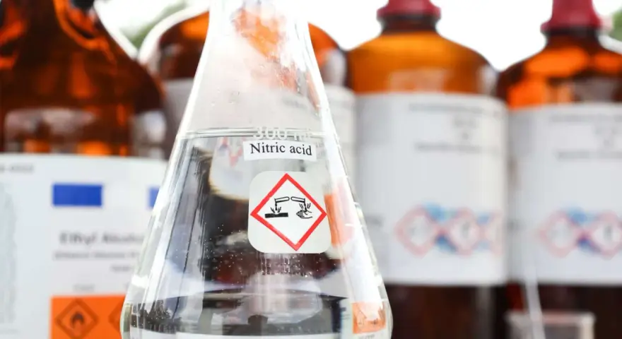 Ein Becherglas mit Salpetersäure (HNO3) und einem Gefahrensymbol. Im Hintergrund stehen weitere braune Glasflaschen mit Chemikalien.