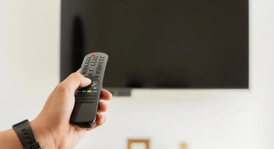 Dłoń trzymająca pilot do telewizora na tle wyłączonego ekranu.