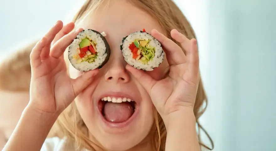 Uśmiechnięta dziewczynka trzyma kawałki sushi dla dzieci przed oczami, udając, że ma rolki zamiast oczu.