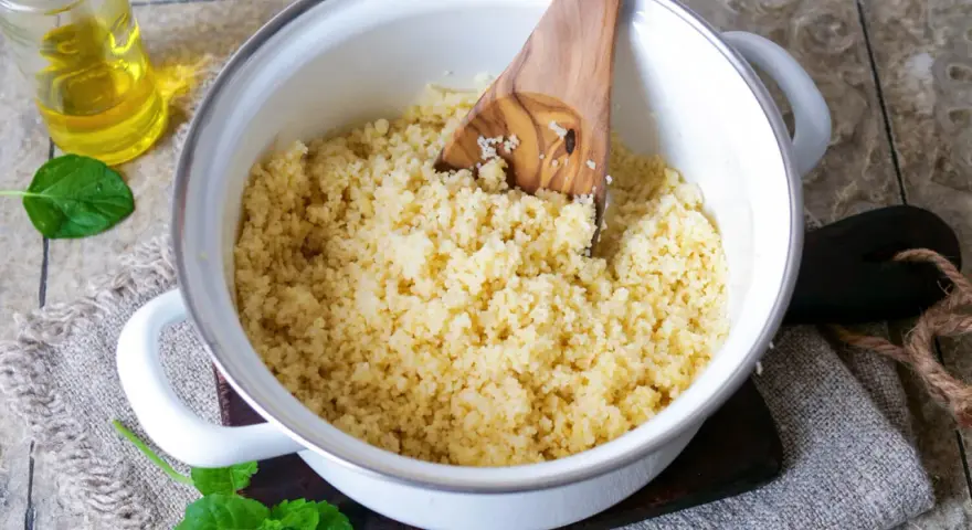 Ein Holzlöffel lockert den frisch zubereiteten Couscous in einem Topf auf. Daneben stehen Öl und Minze.